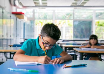 Focused young boy in glasses sitting at desk and writing in copybook in class. Copy space. Education or back to school concept